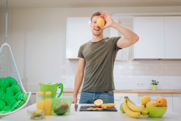 Young smiling handsome man holds sweet orange in front of face while cooking fresh fruits in the...