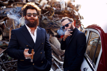 Close up of a bearded mens in black suits and sunglasses smokes cigars near vintage car, on the...