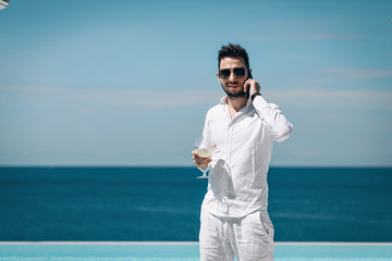 Photo of young man in white shirt standing poolside and talking on mobile phone, drinking mojito and enjoying beautiful blue sea view. Travel and business concept. Phuket. Thailand.