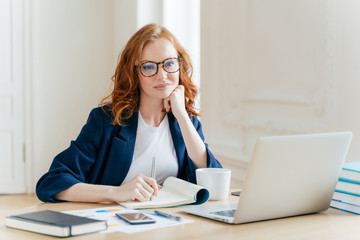 Redhead curly woman office worker analyzes data, makes accounting report, poses in coworking...