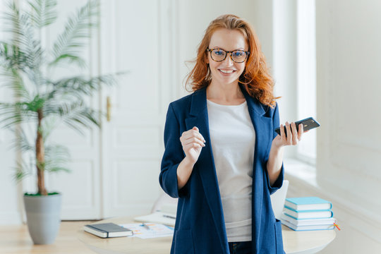 Smiling prosperous ginger female banker makes payment on online servie, waits for important call, holds modern cell phone, looks postively at camera, dressed in elegant clothes, poses at office