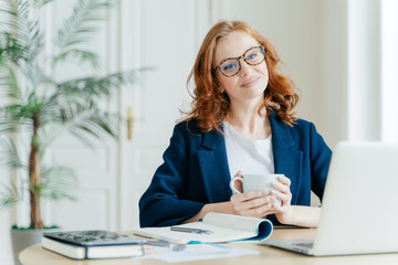 Pleased female teacher makes research work on laptop computer, writes down information in notepad,...