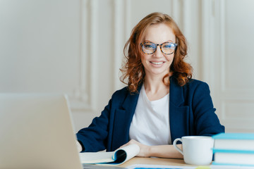 Glad smiling red haired young female concentrated on creating new business project, owns corporation, sits in front of laptop computer, wears optical glasses and elegant outfit, updates software