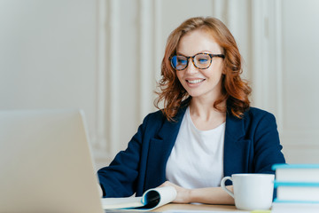 Glad lovely woman has ginger hair, positive smile, sits with laptop computer at desktop, happy to...