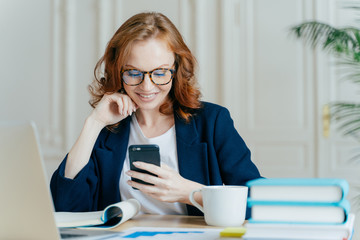 Photo of lovely female sits with smartphone device, types feedback, works in office on up to date...