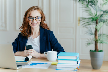 Photo of red haired smiling young woman installs app on mobile phone, checks newsfeed, works on...