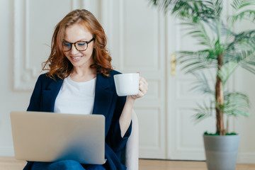Optimistic red haired young successful female freelancer works remotely in coworking space, makes...