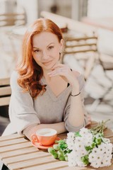 People and lifestyle concept. Red haired female has pleased expression, drinks coffee, poses in cafeteria with flowers, has date with boyfriend, looks directly at camera, enjoys spare time on terrace