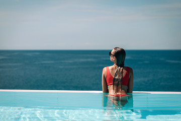Young woman relaxaing in the swimming pool looking at the ocean view in background l. Beautiful seascape. Trip to warm destination. Phuket. Thailand.
