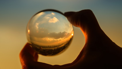 The hand holds glass ball which reflects sunset sky over city.