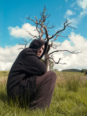 A young man wearing a long dark jacket in front of a sinister dead tree in the middle of a countryside field. Man appears worried and alone. mental health and contemplation.