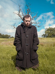 A young man wearing a long dark jacket in front of a sinister dead tree in the middle of a...