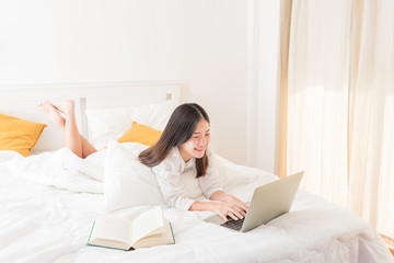 Young woman lying in bed, reading a book and using laptop computer