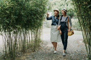 full length asian women having fun together outdoors on hike through amazing bamboo forest trail. young girl pointing finger on tree showing sharing talking with friend. cheerful female tourist kyoto - obrazy, fototapety, plakaty