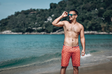 Outdoor fashion portrait of handsome man posing at amazing tropical beach, in nice sunny day, beautiful view on blue sky and ocean, wearing casual orange shorts and sunglasses.