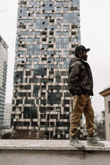 Handsome African American with dreadlocks in brown trousers and a blue sweater in a black cap on his head against the background of houses, city