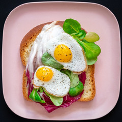 Healthy breakfast sandwiches concept. Bread toasts with fried eggs and green salad on pink plate on black background, top view. Dietary food