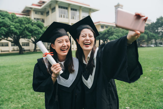 two cheerful asian college girl study abroad standing outdoor on lawn finish bachelor degree. Capturing happy moments. happy female student in graduation gowns making selfie smiling hold smart phone