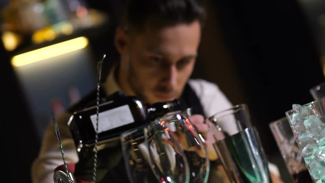 Diagonal view of bar counter with different drink glasses and bartender pouring alcohol from bottle into steel jigger to add in shaker. Alcoholic cocktail preparation in nightclub