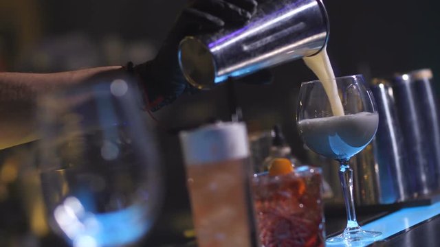 Closeup of bartender hand with steel shaker pouring Bahia cocktail in wineglass standing on bar counter among other prepared drinks, diagonal view. Barman finishing beverages preparation in nightclub