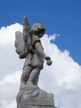 Scene in a cemetery: an old stone statue of a little angel with wings holding a flower. The blue sky with big clouds gives drama to the scene.