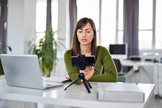 Charming Caucasian Blonde Woman In Green Outfit Adjusting Smart Phone For Video Call. In Front Of Her Laptop, Office Interior.