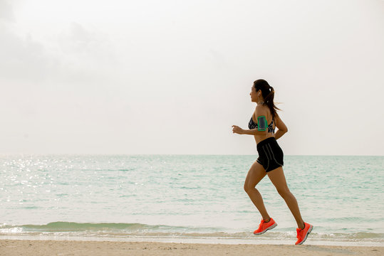 Asia woman in sports bra  listening to music on smartphone armband jogging on the beach. Fitness female  runner running in morning at sea , summer healthy active lifestyle concept
