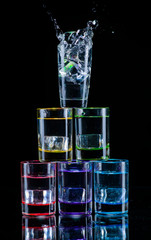 Multicolored glasses filled with alcoholic drinks, with splases of ice cubes falling inside, standing on the mirror surface. Black background. Conceptual, celebrated, commercial design