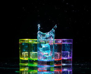 Multicolored glasses filled with alcoholic drinks, with splases of ice cubes falling inside, standing on the mirror surface. Black background. Conceptual, celebrated, commercial design