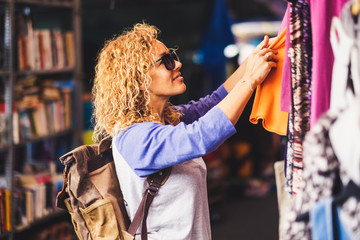Cheerful traveler young blonde curly woman looking and choosing clothes at the used market during...