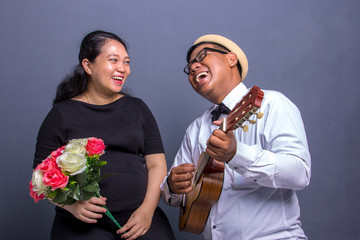 Musical husband entertaining his pregnant wife, singing a song with an ukulele, over gray background