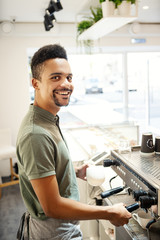 Side view of young positive ethnic bartender smiling at camera while pouring coffee into cup from...