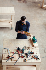 From above young woodworker measuring wooden plank with ruler at workbench covered with tools and...