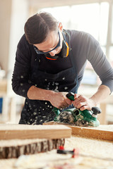 Young woodworker sanding wooden plank with electric flatbed sander, sawdust flying away