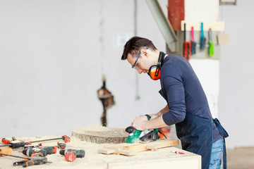 Side view of young woodworker in protective eyeglasses making wooden detail and using electric sanding machine at workbench with equipments and sawdust on in against blurred background