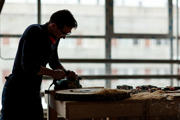 Side view backlit of young woodworker using electric planing machine at workbench covered with sawdust and wood shavings - obrazy, fototapety, plakaty