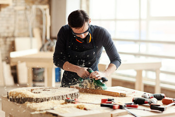 Young woodworker in protective eyeglasses making wooden detail and using electric planing machine at workbench covered with equipments and sawdust on blurred background - obrazy, fototapety, plakaty
