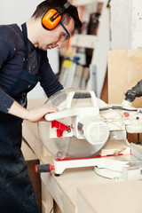 Side view of young woodworker in protective eyeglasses and ear muffs sawing wooden plank with...