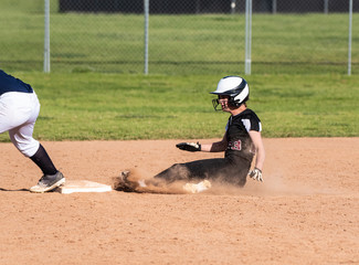 Skilled teenage softball player sliding safely into second base in a cloud of dust.