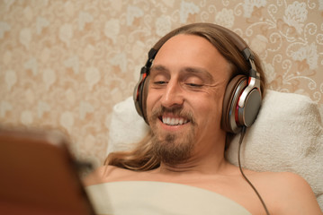 A man lying in bed and listening to music. Portrait of a handsome man from above with headphones in bed. Portrait of a guy with a mustache and beard.