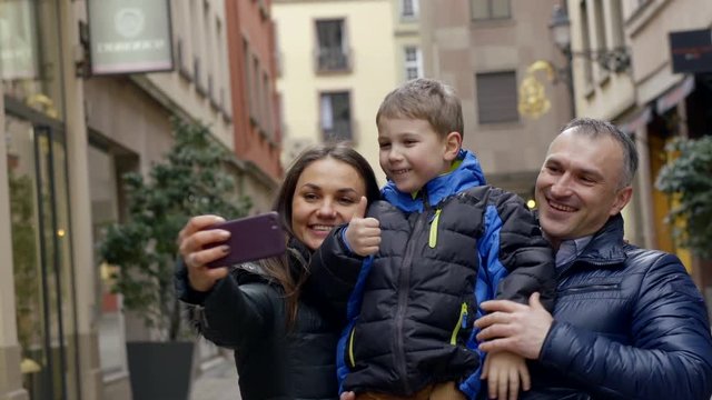 The family - mom, dad and son are on the background of city houses. They hold their son in their arms and take pictures on the phone, take selfies.