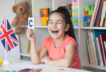 Learning English. Happy cute child girl sitting at a table and draws with a white bookcase, colored books and toys and British flag behind. Back to school and happy time! 