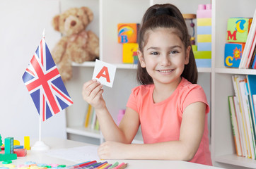 Learning English. Happy cute child girl sitting at a table and draws with a white bookcase, colored books and toys and British flag behind. Back to school and happy time! 
