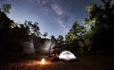 Young couple hikers man and woman having a rest beside camp, bonfire and glowing tourist tent at night. On background beautiful night starry sky full of stars and Milky way, mountain rocks, trees