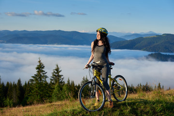 Athlete female cyclist with yellow bicycle in the mountains, wearing helmet, enjoying valley view on sunny morning. Foggy mountains, forests on the blurred background. Outdoor lifestyle activity