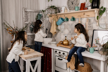 Three charming little girls in white sweaters and blue jeans play on an old-fashioned kitchen