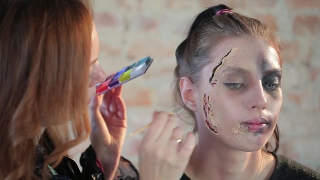Close-up woman professional makeup artist makes wax plastic makeup in form of bloody wound for art cinema and paints face of cute girl and creates image for shooting scene on background brick wall