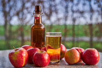 Close up picture of bottle and glass with fresh sparkling apple cider and red riped apples on stone granit table in garden restaurant during spring sunny evening taken in golden hour before sunset.