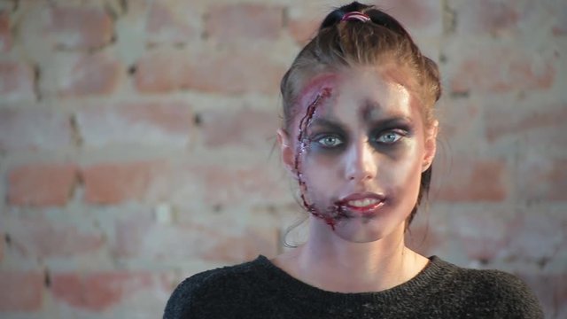 Close-up cute girl with beautiful blue big eyes whose face has wax plastic makeup in form of torn bloody wound, she looks at camera and shows makeup on blurred background of red brick wall