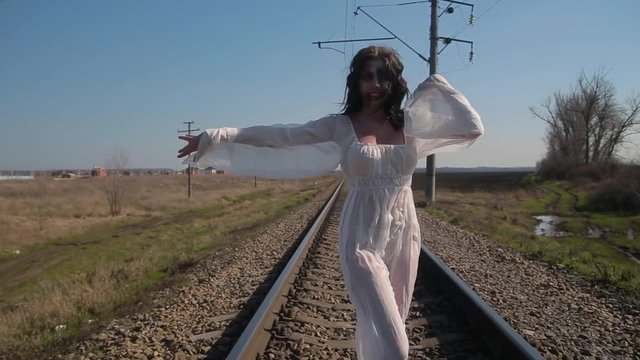 Camera motion, amazing slim unusual girl in white translucent dress and with art plastic wax makeup on her face in form of torn bloody wound makes unusual dance moves on railway tracks in sunny day